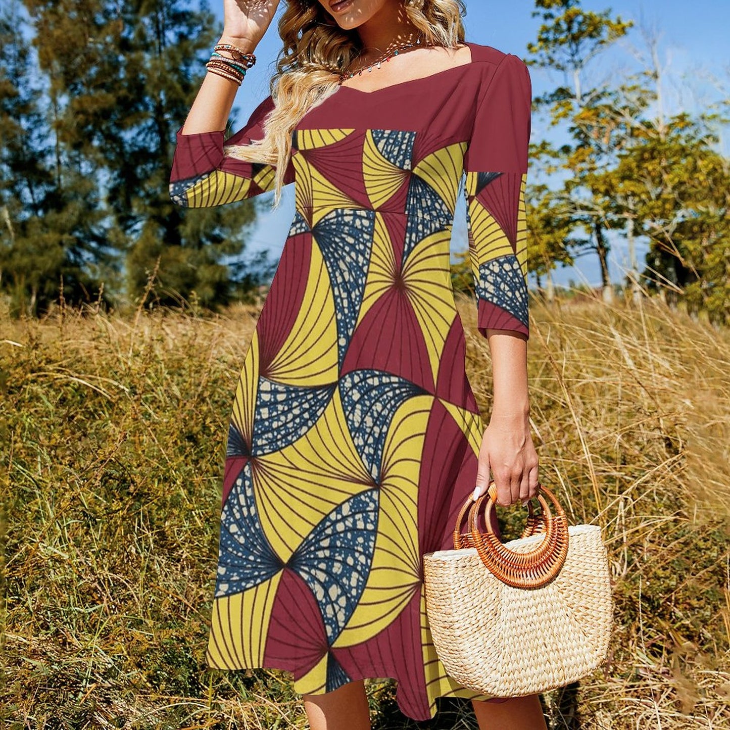 A woman outdoors in a field wearing a loose, three-quarter sleeve dress with a bold geometric fan pattern in maroon, mustard yellow, and navy blue. She is holding a woven straw handbag with a round wooden handle.