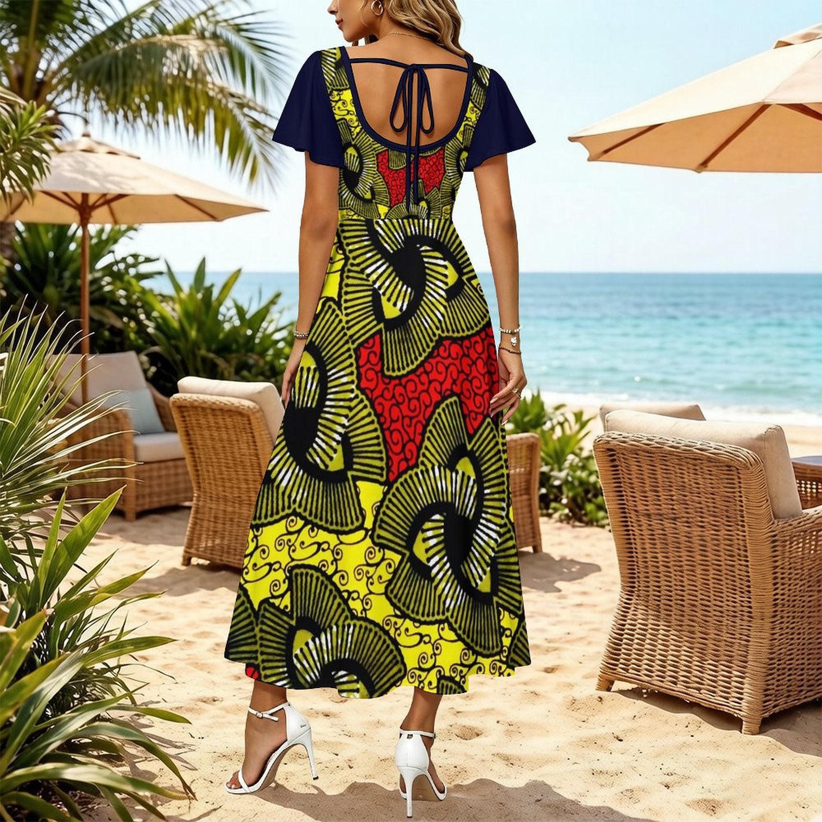 A woman stands on a sandy beach wearing a short-sleeve maxi dress with a bold African print. The dress features a navy blue back panel and sleeves, with a body pattern of yellow, green, black, and red floral motifs.