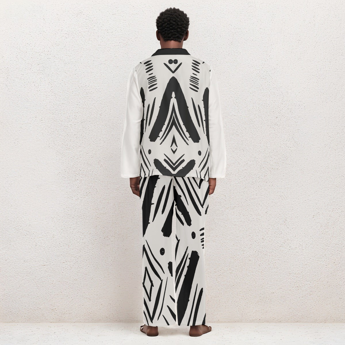 A model viewed from the back wearing a matching long-sleeve white and black geometric patterned robe and trousers set, standing against a textured off-white wall.