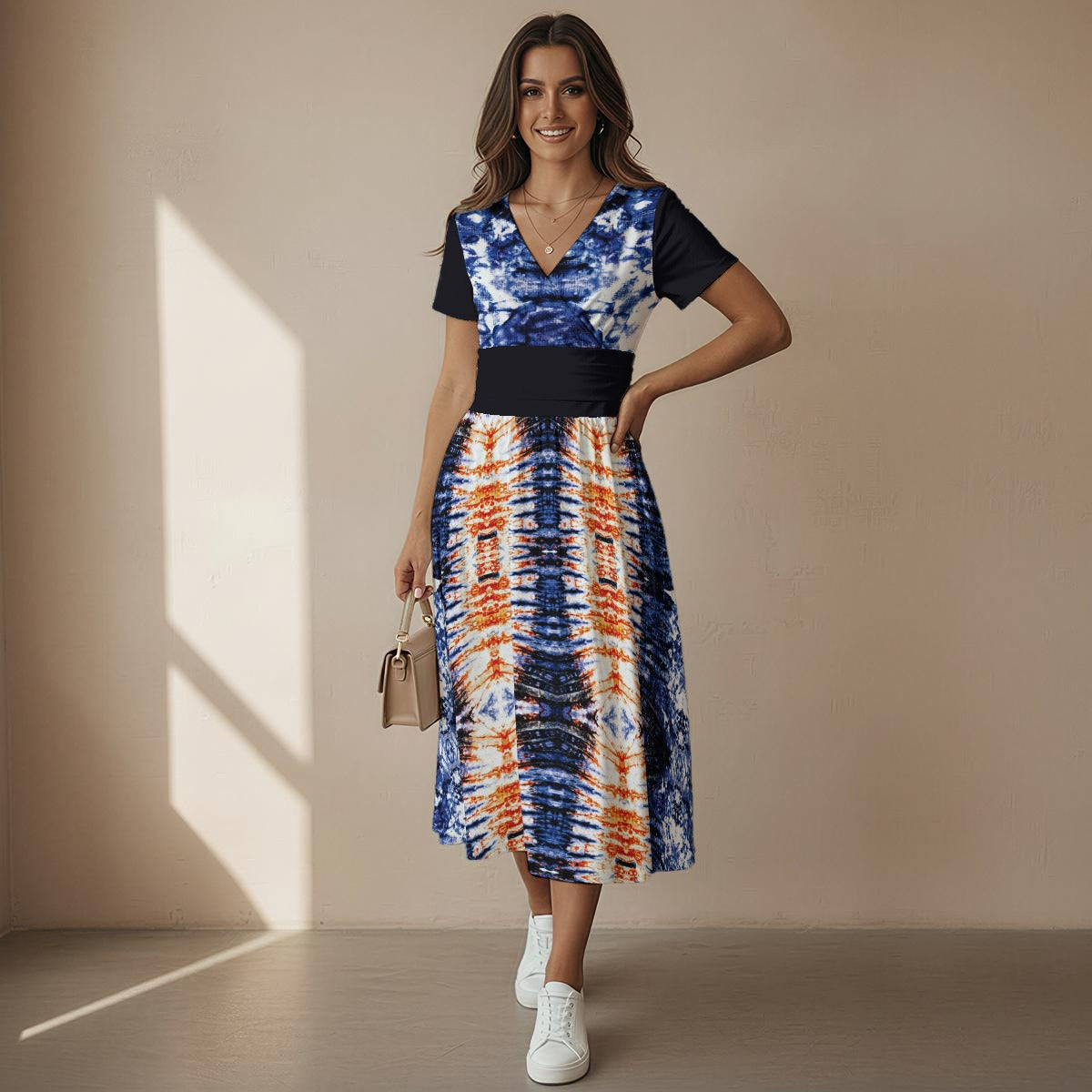 A woman models a short-sleeve midi dress with a navy blue and white abstract pattern on the bodice and a multicolor orange, blue, and white ikat-style patterned skirt, paired with white sneakers and a small beige handbag against a beige wall with window shadows.