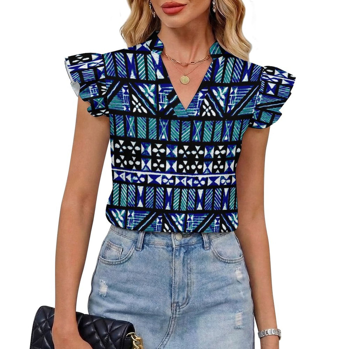 A woman wearing a short-sleeve V-neck blouse with a bold geometric tribal pattern in blue, black, and teal, paired with light-wash distressed denim shorts and holding a black quilted clutch.