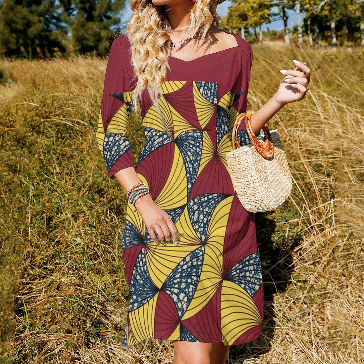 A woman wearing a three-quarter sleeve dress with a bold geometric pattern in maroon, yellow, and navy blue, standing in a field of dry grass and holding a woven straw handbag.