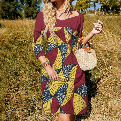 A woman wearing a three-quarter sleeve dress with a bold geometric pattern in maroon, yellow, and navy blue, standing in a field of dry grass and holding a woven straw handbag.