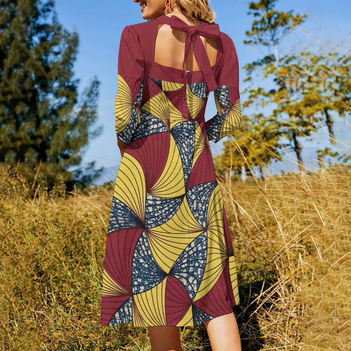 A woman wearing a long-sleeve midi dress with a geometric African print in maroon, mustard yellow, and navy blue, photographed from behind in an outdoor grassy field with trees and blue sky.