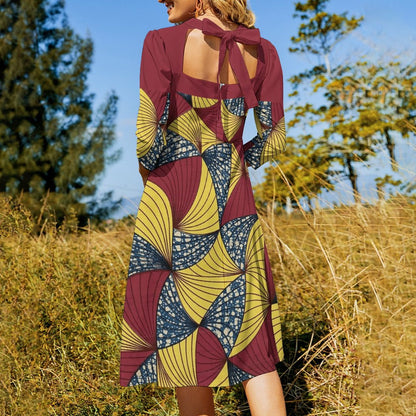 A woman wearing a long-sleeve midi dress with a geometric African print in maroon, mustard yellow, and navy blue, photographed from behind in an outdoor grassy field with trees and blue sky.