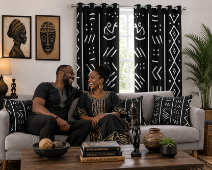 A couple sits on a light gray sofa in a styled living room, with black curtains featuring white geometric and tribal patterns hanging behind them. The room includes framed wall art, a lamp, a plant, and a wooden coffee table in the foreground.